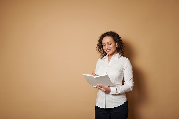 Femme souriante debout sur un fond beige et prendre des notes en lisant un livre