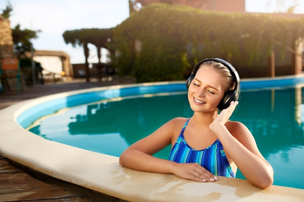 Femme souriante détendue écoutant de la musique dans des écouteurs se baignant dans la piscine fille blonde aime