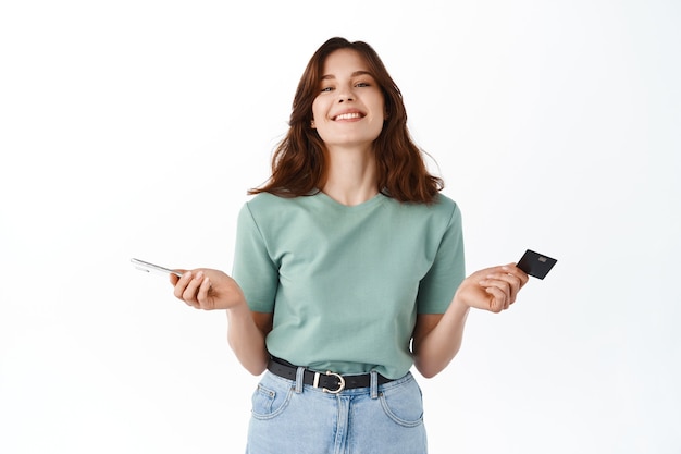 Femme souriante et détendue, facile à payer avec une carte de crédit et un téléphone portable, ne semble pas dérangée devant le visage heureux après avoir envoyé de l'argent avec une application pour smartphone, faire des achats en ligne, mur blanc