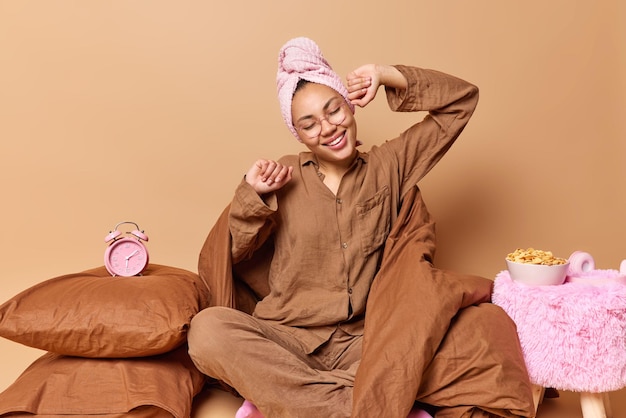 Une femme souriante et détendue positive se réveille tôt le matin après avoir dormi porte un pyjama et une serviette de bain enroulée sur la tête pose sur une couette douce réveil sur un oreiller et un bol de céréales près