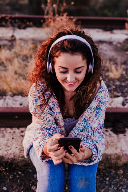 Une femme souriante écoutant de la musique et utilisant un téléphone alors qu'elle est assise sur la voie ferrée