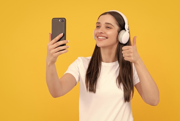 Femme souriante écouter de la musique dans les écouteurs avec smartphone montrer le pouce vers le haut sur fond jaune selfie