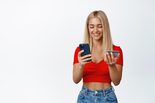 Femme souriante élégante payant la commande en ligne sur smartphone avec carte de crédit à la main, debout sur fond blanc