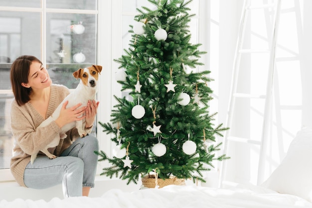 Une femme souriante embrasse un animal de compagnie avec amour a une bonne humeur pose ensemble près d'une belle échelle de murs blancs de sapin de Noël décoré et d'une fenêtre autour de passer du temps à la maison Vacances d'hiver