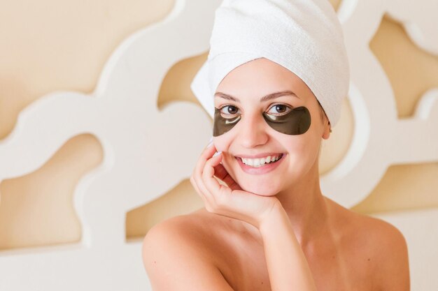 Femme souriante enveloppée dans une serviette après le bain et avec des taches noires sous les yeux pose sur le lit Concept de traitement de beauté