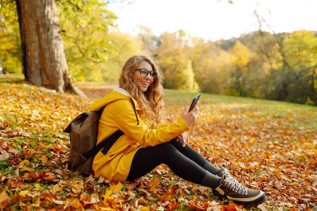 Une femme souriante est assise dehors sur la pelouse parmi les feuilles jaunes d'automne avec un téléphone dans les mains