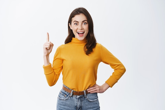 Femme souriante et excitée montrant une publicité, pointant le doigt vers le haut et regardant amusé devant, debout contre un mur blanc