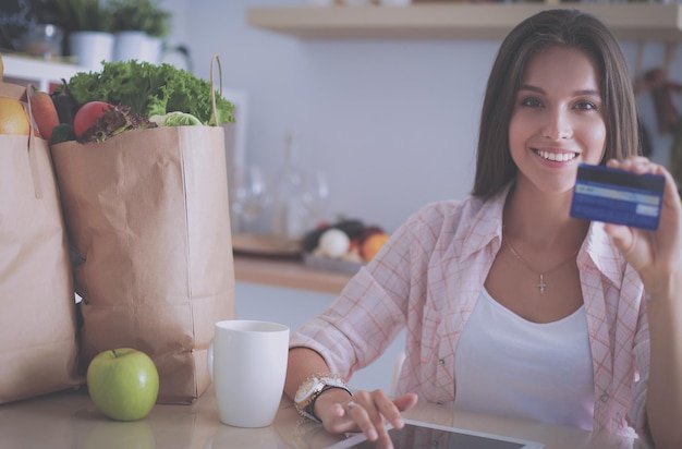 Femme souriante faisant des achats en ligne à l'aide d'une tablette et d'une carte de crédit dans la cuisine