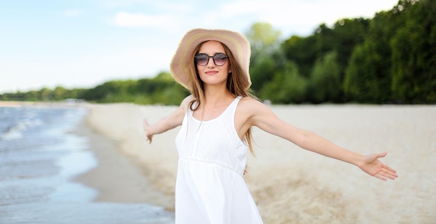 Femme souriante heureuse dans le bonheur libre sur la plage de l'océan debout avec un chapeau, des lunettes de soleil et des mains ouvertes. Portrait d'un modèle féminin multiculturel en robe blanche d'été appréciant la nature