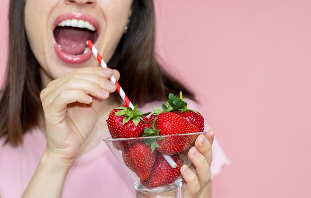 femme souriante heureuse tenant un bol en verre avec des fraises fraîches à la main et une paille à boire dans la bouche