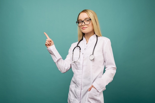 Femme souriante jeune médecin avec stéthoscope pointant sur copyspase. Isolé sur l'espace bleu.