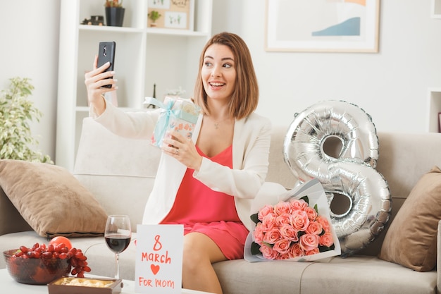 Femme souriante le jour de la femme heureuse tenant un cadeau et prenant un selfie assis sur un canapé dans le salon