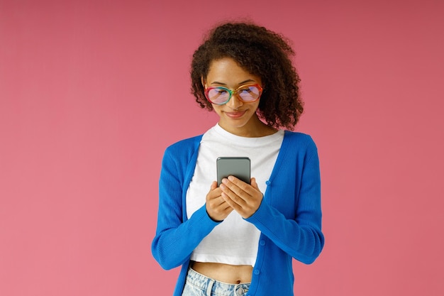 Une femme souriante en lunettes regardant son téléphone portable alors qu'elle se tient sur un fond de studio rose