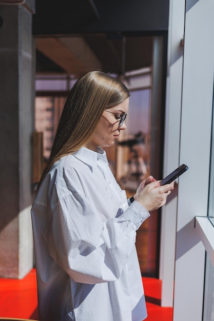 Femme souriante à lunettes en tenue décontractée regardant une vidéo positive sur un téléphone connecté au wifi une blogueuse en chemise blanche contre une grande fenêtre profitant de la détente pendant le travail à distance