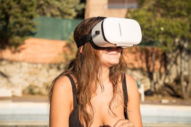 Femme souriante en maillot de bain à l'aide de lunettes de réalité virtuelle assise au bord de la piscine