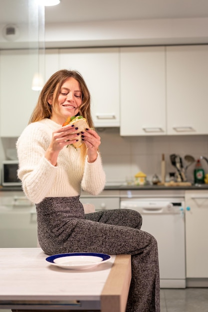 Femme souriante mangeant un sandwich aux légumes dans la cuisine à la maison une alimentation saine