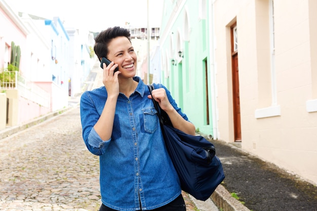 Femme souriante marchant dans la rue avec téléphone portable et sac