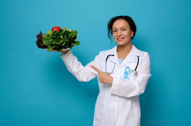 Femme souriante, médecin nutritionniste en blouse médicale blanche avec des points de ruban de sensibilisation bleus sur une assiette pleine de nourriture végétalienne crue saine. Concept de la Journée mondiale du diabète sur fond coloré avec un espace pour l'annonce
