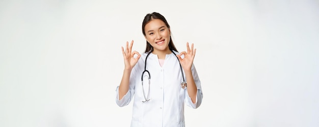 Femme souriante médecin travailleur de la santé asiatique en uniforme de clinique médicale montre des signes corrects approuver qch donner la permission au patient se tient sur fond blanc