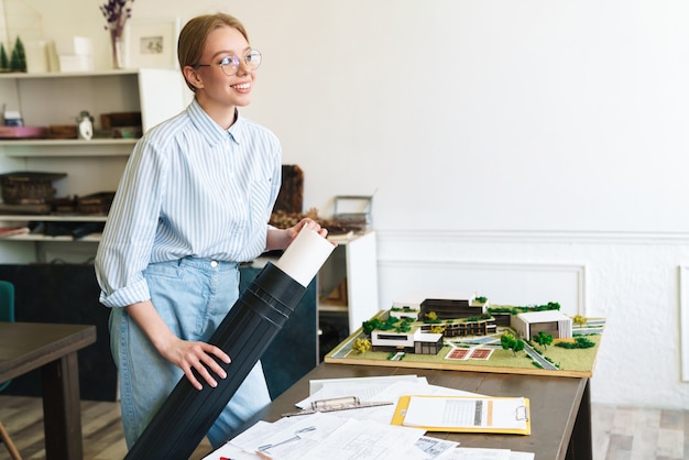 femme souriante et mignonne architecte à lunettes travaillant avec des dessins lors de la conception d'un projet sur le lieu de travail