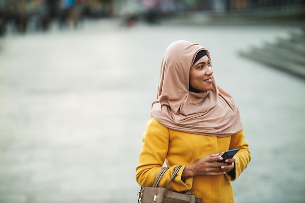 Photo femme souriante musulmane noire portant le hijab debout dans un environnement urbain et utilisant son smartphone.