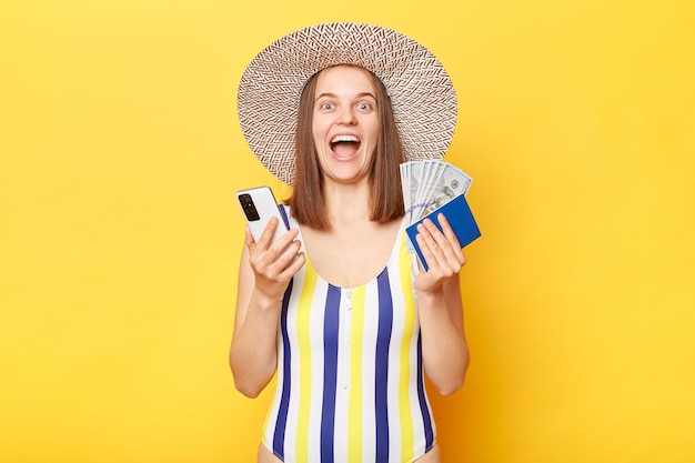Femme souriante optimiste sur la station vêtue d'un maillot de bain rayé et d'un chapeau de paille posant isolé sur fond jaune tenant son passeport et utilisant un téléphone intelligent criant avec un visage heureux