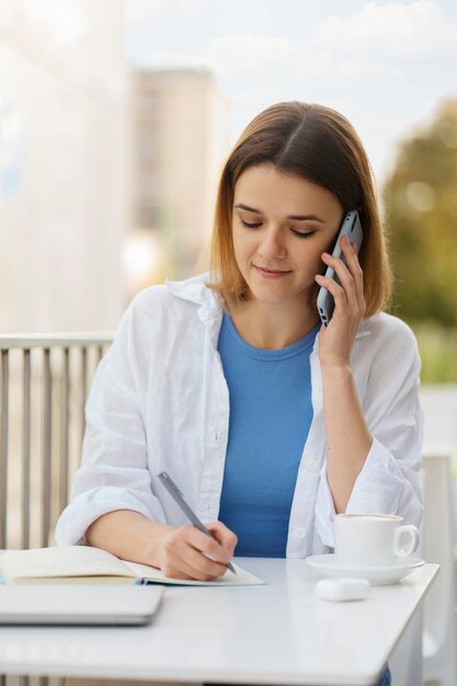 Femme souriante parlant au téléphone mobile en prenant des notes projet de travail concept technologique