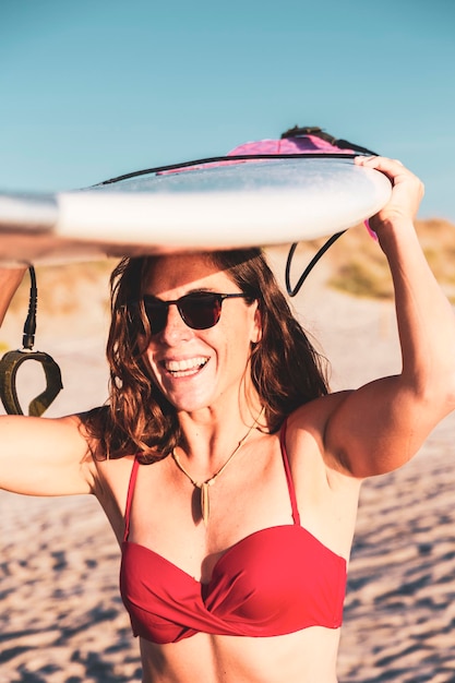 Une femme souriante sur la plage porte une planche de surf reposant sur sa tête