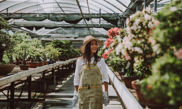 Femme souriante portant un chapeau debout au milieu des plantes en pot dans la serre