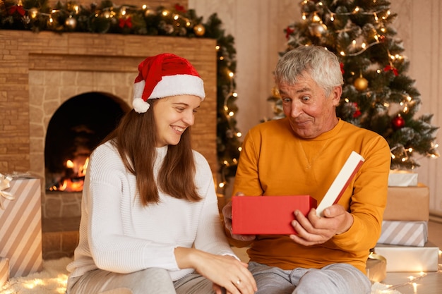 Femme souriante portant un pull blanc et un chapeau de père Noël assis avec son père dans le salon avec des décorations de Noël, célébrant le nouvel an ensemble.