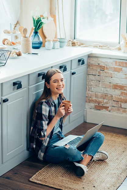 Femme souriante positive assise sur le sol de la cuisine avec une tasse et un appareil moderne