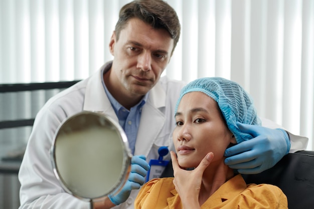 Une femme souriante qui se regarde dans le miroir et apprécie le résultat des injections de remplissage.