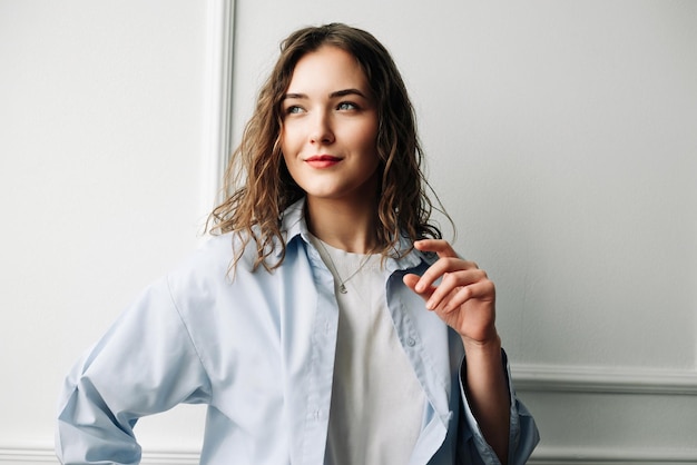 Une femme souriante et réfléchie aux yeux tristes dans une chemise décontractée bleue pose sur fond de mur blanc à la maison regarde ailleurs en se souvenant des souvenirs doux-amers de son ex-petit ami jouant avec ses cheveux