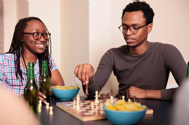 Femme souriante regardant un ami pensif déplaçant une pièce d'échecs à bord. Les gens occasionnels se détendent en jouant à des jeux de stratégie tout en profitant de loisirs amusants ensemble à la maison dans le salon.