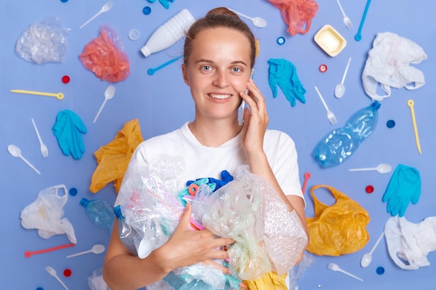 Femme souriante et satisfaite portant une chemise blanche isolée sur un mur bleu avec des ordures autour tenant des déchets parlant sur un téléphone portable en regardant la caméra sauver la terre nettoyer la planète