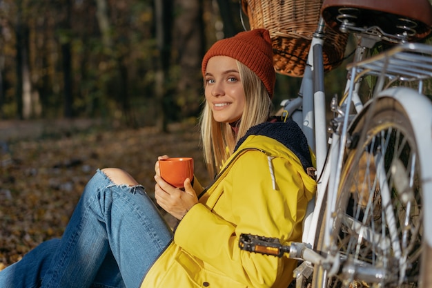 Femme souriante se détendre dans la forêt d'automne, tenant une tasse de café