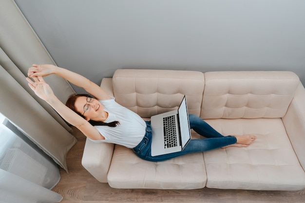 Une femme souriante se repose avec un ordinateur portable après le travail effectué sur un canapé moelleux à la maisonconcept de stressfr...