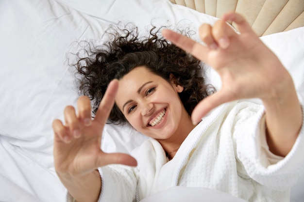 Femme souriante se réveillant dans son lit et étirant ses bras en faisant un cadre photo avec les mains faisant semblant
