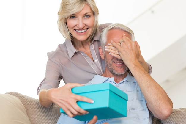 Femme souriante surprenant homme mature avec un cadeau sur le canapé