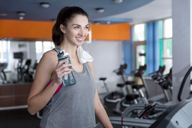 Femme souriante sur tapis roulant avec une bouteille d'eau et une serviette à la salle de sport Belle jeune brune en vêtements de sport Activité sport et mode de vie sain Vertical