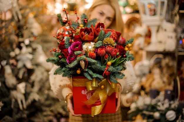 Femme souriante tenant une boîte rouge avec ruban d'or avec différentes fleurs lumineuses décorées de branches de sapin