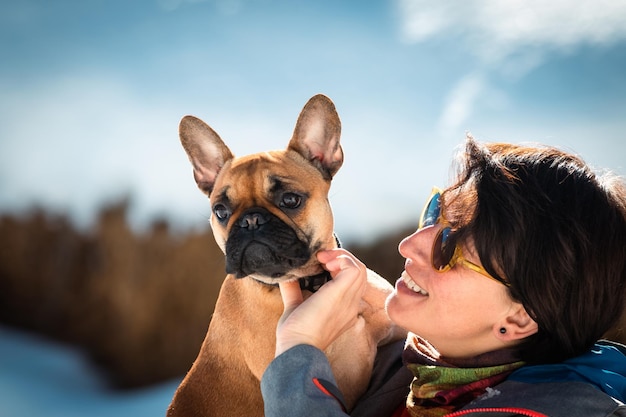 Femme souriante tenant sur les mains le buldog français et jouant avec sur le fond blanc neigeux