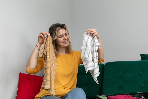 Photo femme souriante tenir dans les mains des t-shirts rayés assis sur un canapé à la maison choix alternatif plier les vêtements