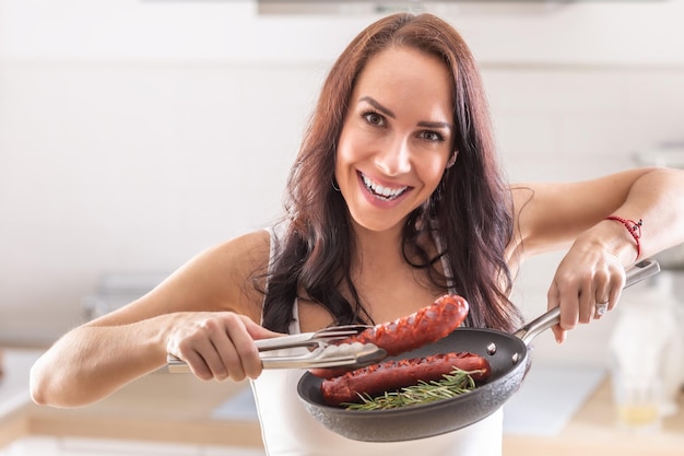 Une femme souriante tient des saucisses fraîchement grillées dans des pinces dans sa cuisine à domicile