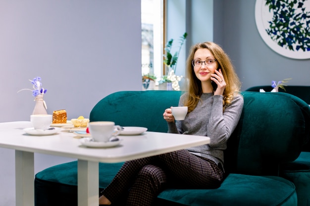 Femme souriante tout en parlant sur son téléphone et manger du gâteau et boire du café au café à l'intérieur, en attendant ses amis