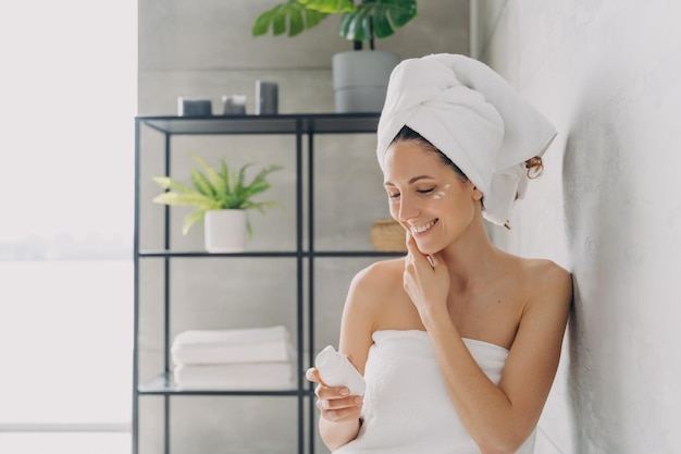 Femme souriante utilisant une crème hydratante pour le traitement du visage dans la salle de bain