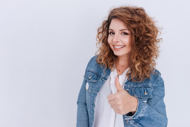 Femme souriante en veste en jean montrant le pouce vers le haut sur un mur gris