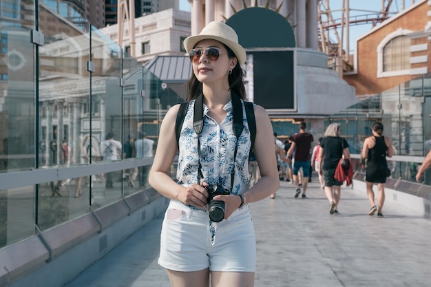 femme souriante voyageuse au chapeau tenant un appareil photo vintage dans la ville moderne de las vegas aux états-unis. Photographe touristique hipster fille marchant à l'extérieur sur un passage piéton. Concept de vacances d'été de voyage.