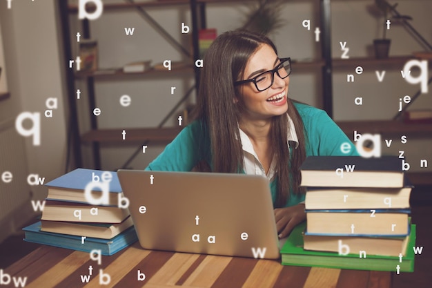 La femme sourit Elle travaille avec beaucoup de livres et un ordinateur portable à la table en bois