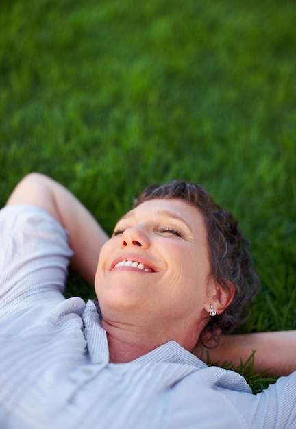 La femme sourit et se détend sur l'herbe pour le confort, la paix et la santé mentale positive, une personne calme, mature et heureuse dans un parc naturel pour les voyages d'été, la liberté et l'environnement sans stress pour le bien-être.